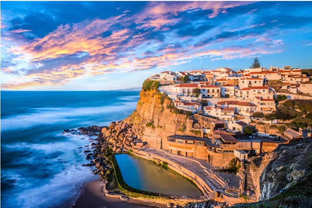 The view of the white houses in Azenhas Do Mar.  They are perched on the cliff edge and below them is a natural swimming pool and beach.