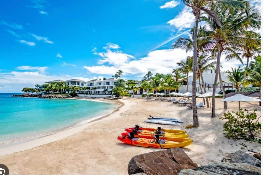 A resort on a golden sand beach. On the beach are two orange and red kayaks with palm tress next to them. This is Hodges Bay Resort the newest Antigua all inclusive family resorts.