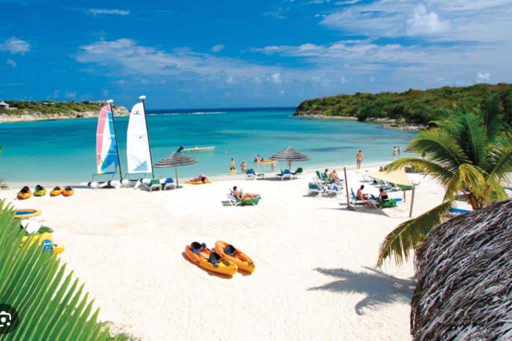 A white snd beach with two red and orange kayaks on it, two sailboats and some sun loungers.