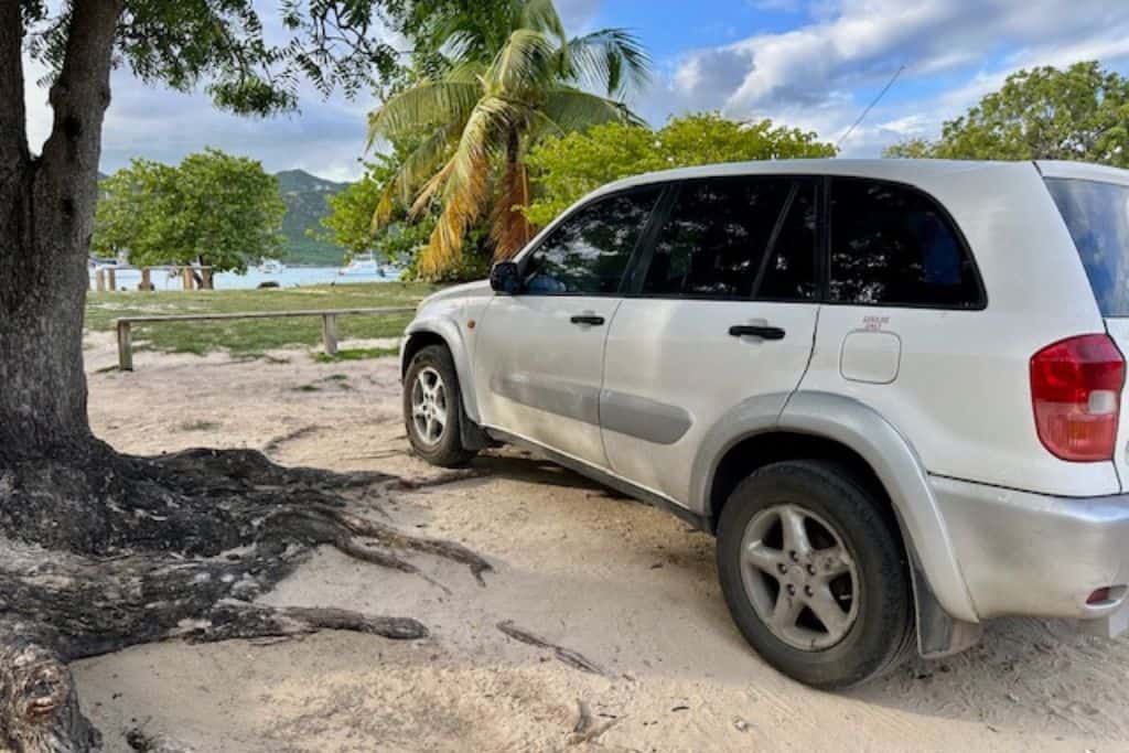 A white Toyota Rav4 rental car in Antigua parked on the beach.  Between the car and the beach is some grass and some palm trees.