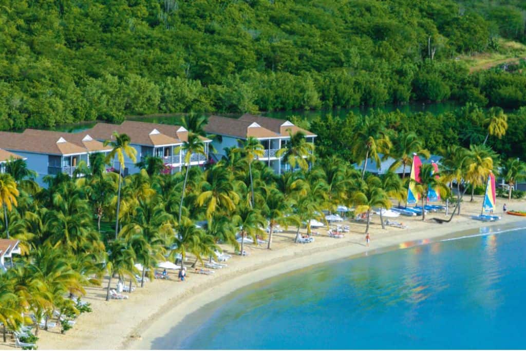 Lush green rain forest surrounds a hotel. In from of rainforest is a beach and on the beach are parasols, loungers and sailboats. This is Carlisle Bay Resort which is one of the best antigua all inclusive family resorts.