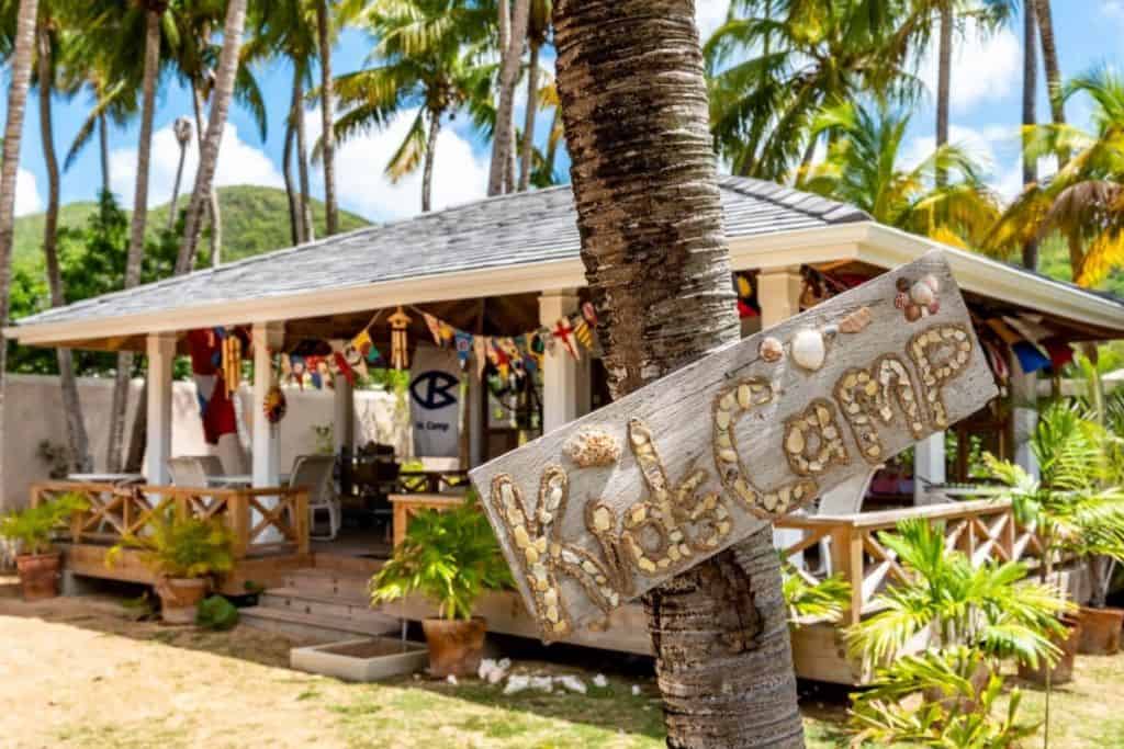 A sign which says kids camp is hung up outside a wooden building surrounded by palm trees. This is Curtain Bluff Resort in Antigua.