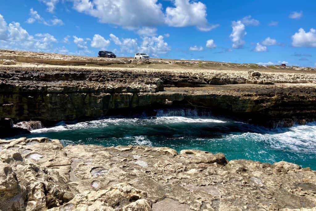 A stone arch with the Atlantic waves crashing on it at Devil's Bridge in Antigua.  On the grass next to the stone arch are two cars.