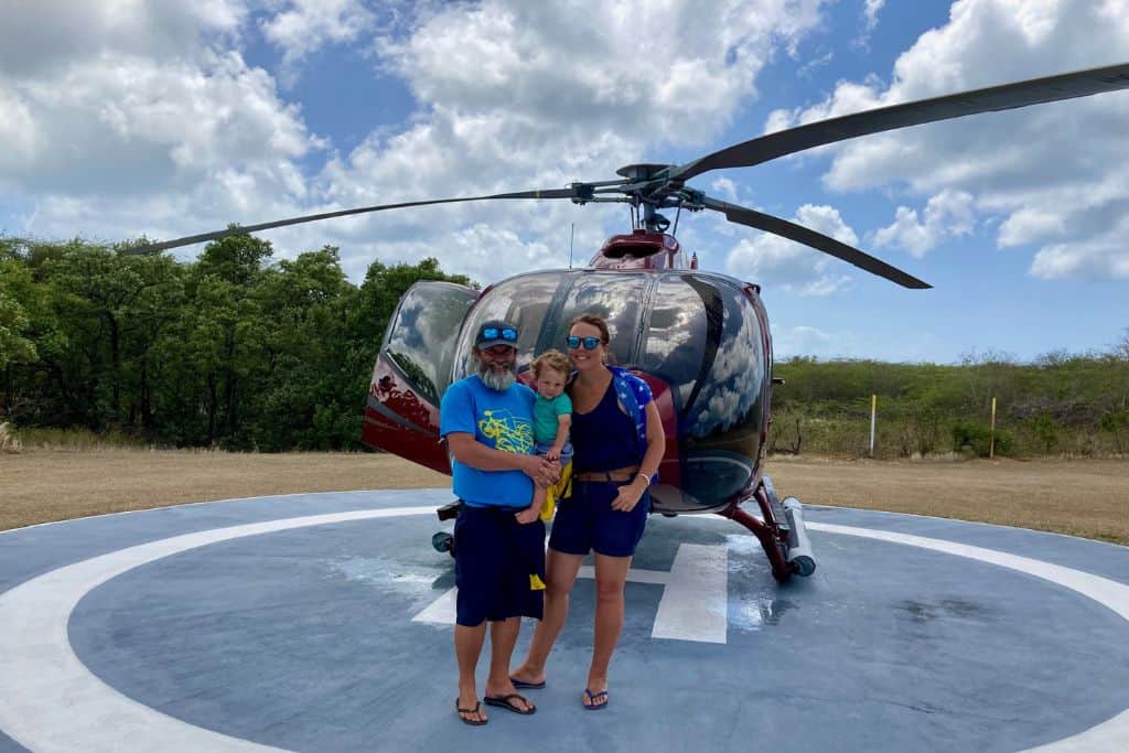 A mum, dad and their toddler son are stood in front of a helicopter which is an amazing thing to do in Antigua.  They are in summer clothes and looking at the camera as the photo is taken.
