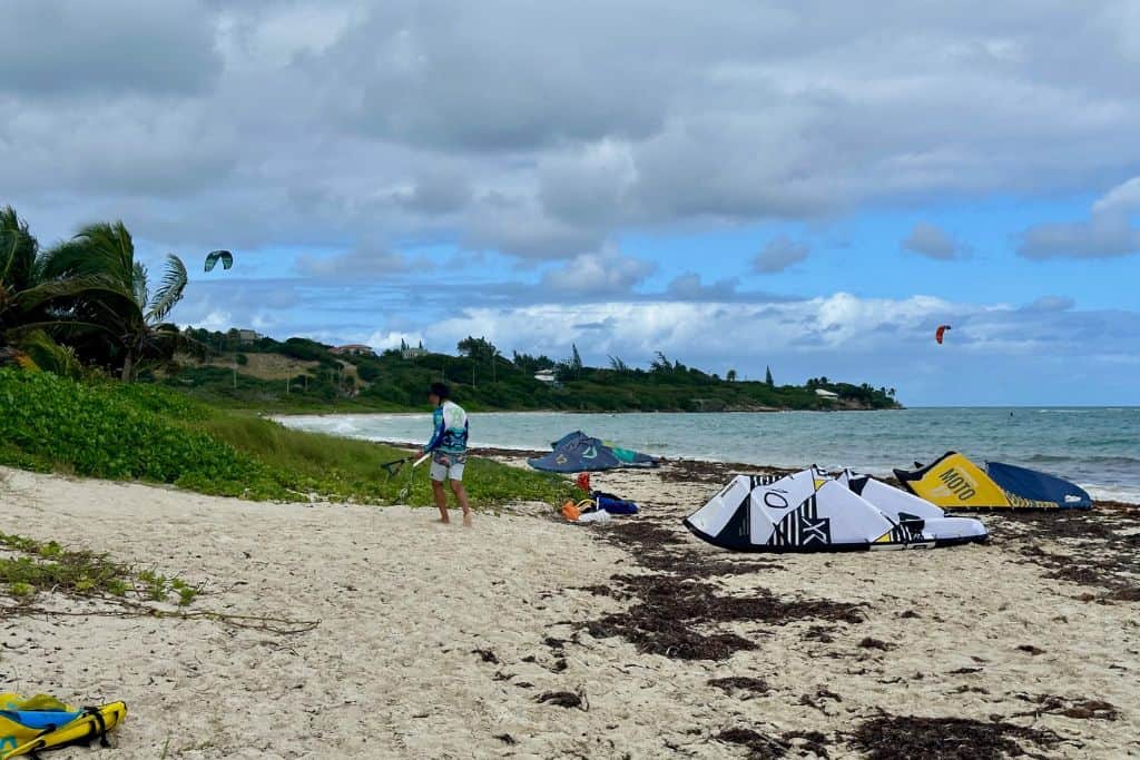Jabberwock beach in Antigua with lots of people kitesurfing in the water and kites on the beach. This is one of the best things to try to do in Antigua.  There is a man also on the beach setting up his kite ready to go out. 