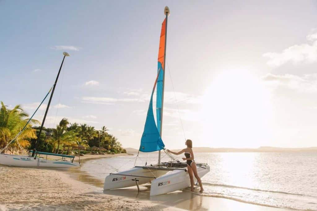 catamaran sil boat on the beach with a woman in a swimsuit stood next to it.  This is Jumby Bay inAntigua.