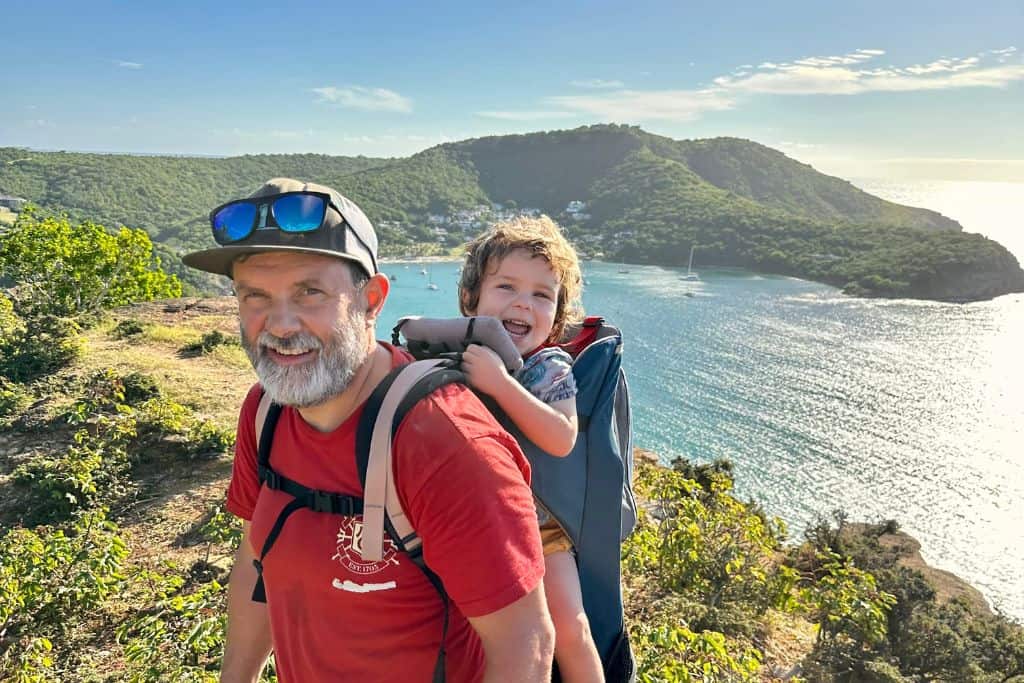 Father in a red t-shirt with a baseball cap is standing with his son in a kids carrier on his back. They are both hiking in Antigua above English Harbour and Nelson's Dockyard. In the background across the entry to English Harbour is the green hill that takes you up to Shirley Heights. This hike called the Goats Trail is one of the best things to do in Antigua