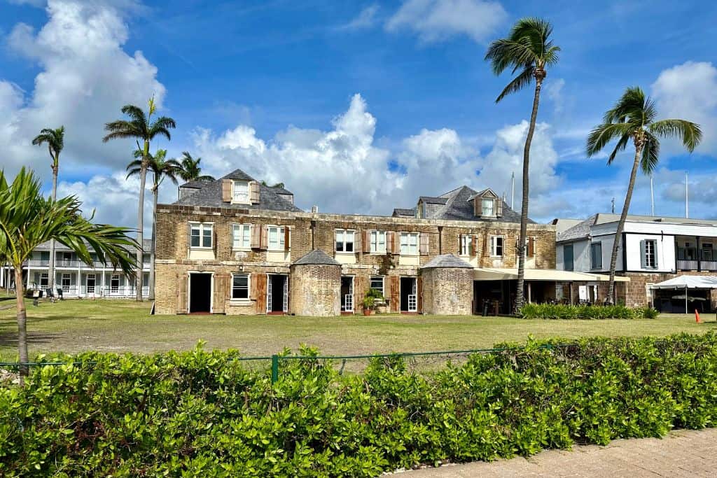 Some of the buildings in Nelson's Dockyard in Antigua.  One of them is the Cooper and Lumber Hotel and has a hedge in front of it and in the middles of the grass in front of the hotel is a palm tree.