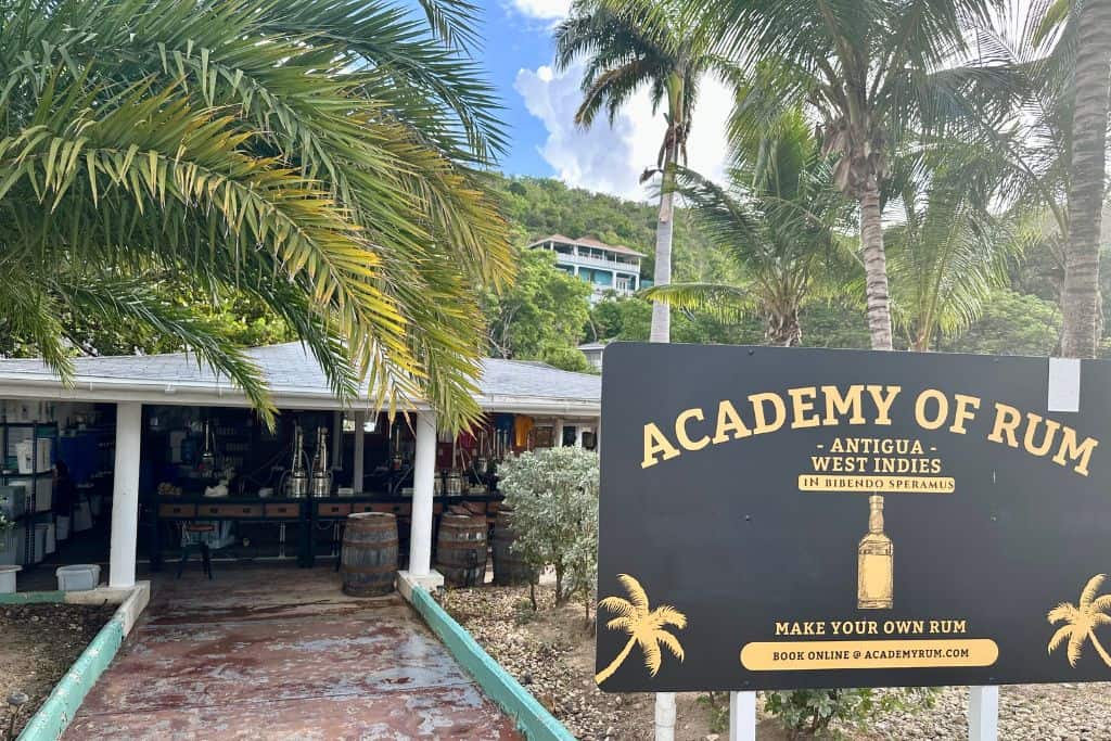 The outside of the rum making academy at Galleon Beach in Antigua.  The building is a wooden on that is open on three sides.  In the building you can see the stills that they use to make the rum, and outside of the building is a sign saying "Academy of Rum" 