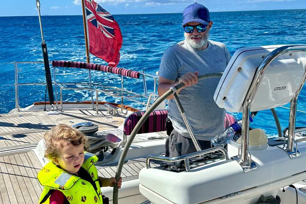 A father in a grey t-shirt and baseball cap with sunglasses is with his son in a yellow life jacket sailing in Antigua with the father at the helm of a sailing  yacht. Sailing is one of the best things to do in Antigua.  Behind them is a British flag that is flying at the back of the boat.