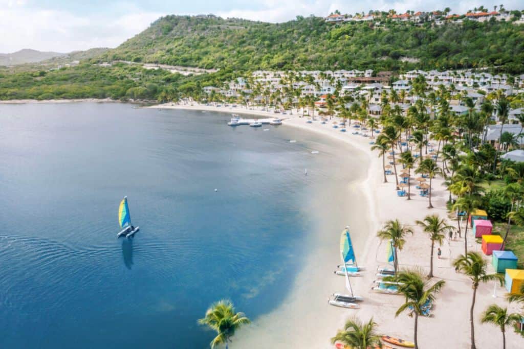 A white sand beach with palm trees on it and a sail boat on the beach. And another sail boat sailing in the water being. This is St. James Club which is one of the best antigua all inclusive family resorts.