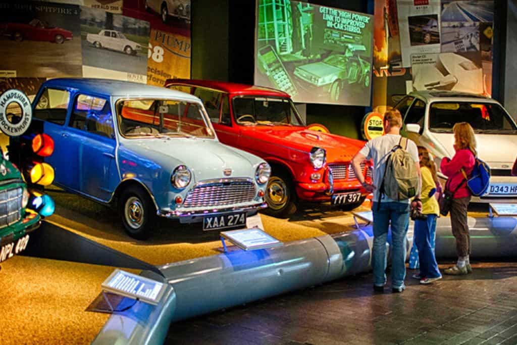 A car display featuring a light blue vintage car and a red vintage car. There is an information screen in the background and two people in the foreground looking at the cars wearing backpacks.  This is Beaulieu National Motor Museum one of the best childrens attractions in hampshire