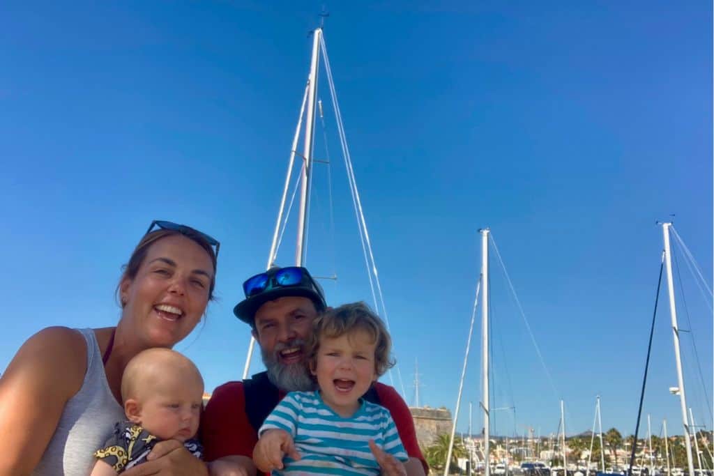 Family of a mum, dad, toddler and baby are in front of some yachts in Caiscais in March which is near Lisbon.