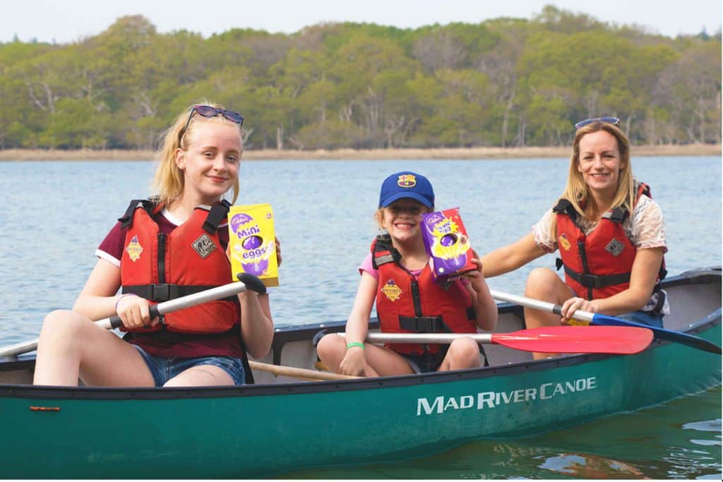 A lady and two girls in an open green canoe on a river with paddles in one hand.  In the other hand the girls each have a Cadburys Cream Egg Easter Egg.  The photo has been used on the New Forest Activities Centre in Hampshire webpage to show that they are offering a canoe Easter Egg hunt.
