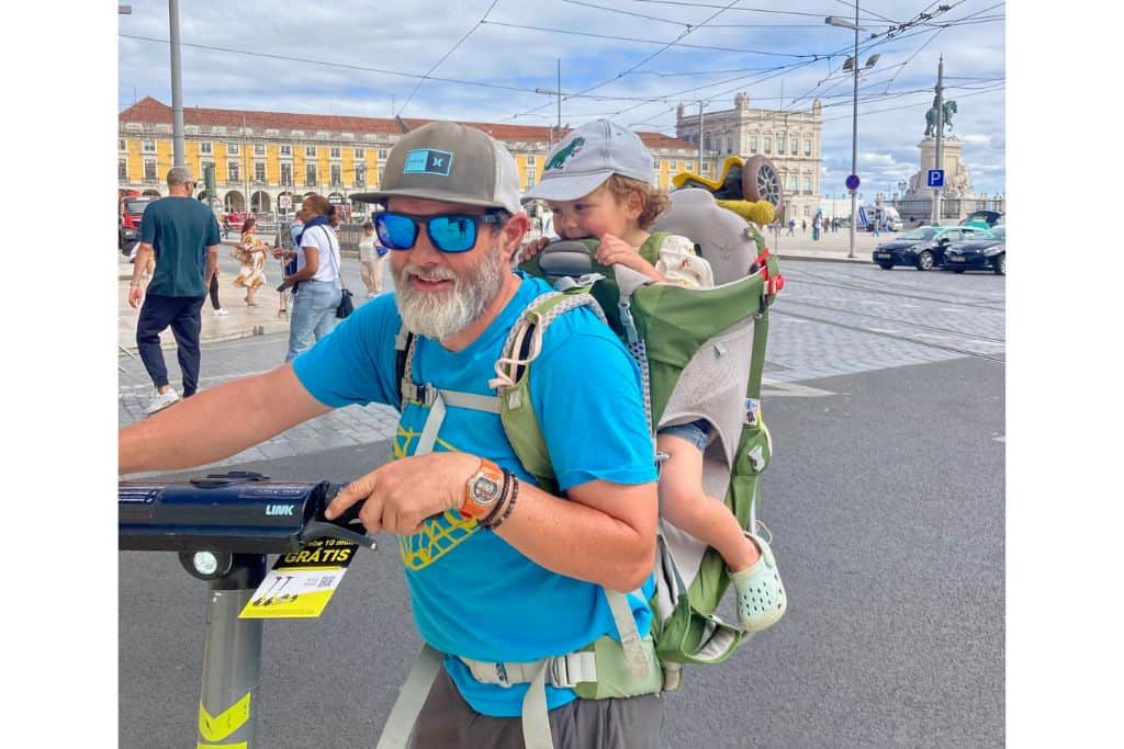 A dad in a bright blue t-shirt with his 3 year old son on his back in green backpack style carrier.  The dad is holding the handle bars of an e-scooter which they are about to use in Lisbon in March.