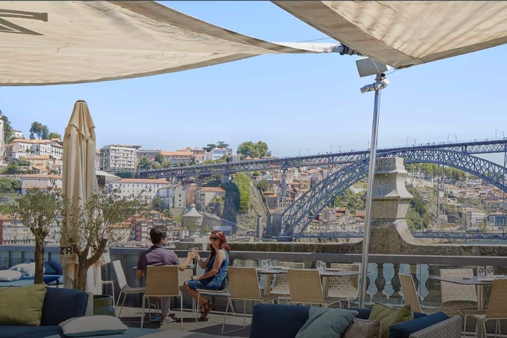The view from the a terrace over one of the many bridges in Porto.  On the terrace are people sat at tables with a sun shade enjoying glasses of port. The terraces is at Espaco Porto Cruz which is one of the best wineries in Porto Portugal.