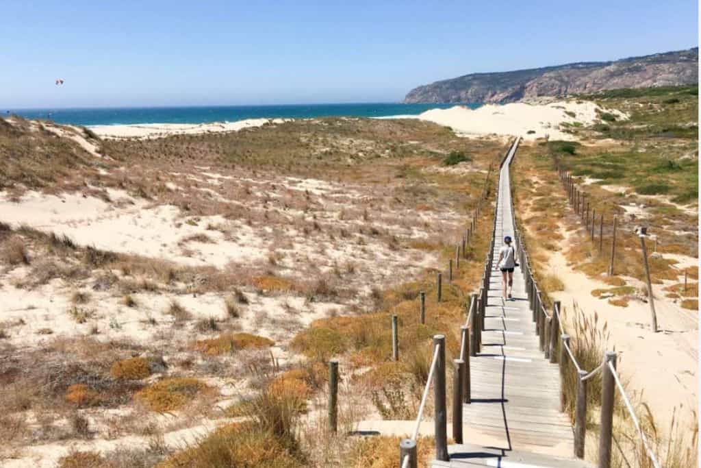 There are white sand dunes with tufts of grass sticking out of them.  Through the dunes is a wooden broadwalk.  The beach is called Praia Guincho and it is near to Lisbon and a fun thing to do with kids.