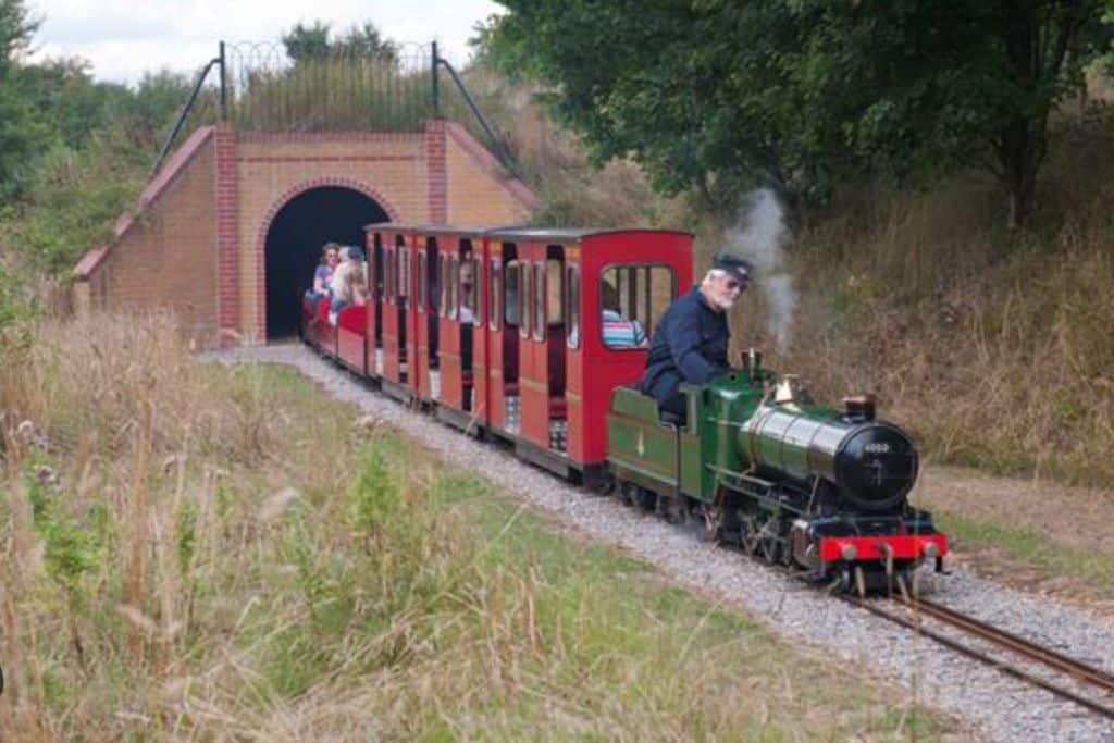 Green steam train with  the driver in the front in a flat cap.  Behind are red carriages with people in them.  The train has steam coming out of it and is coming out of a red brick tunnel.