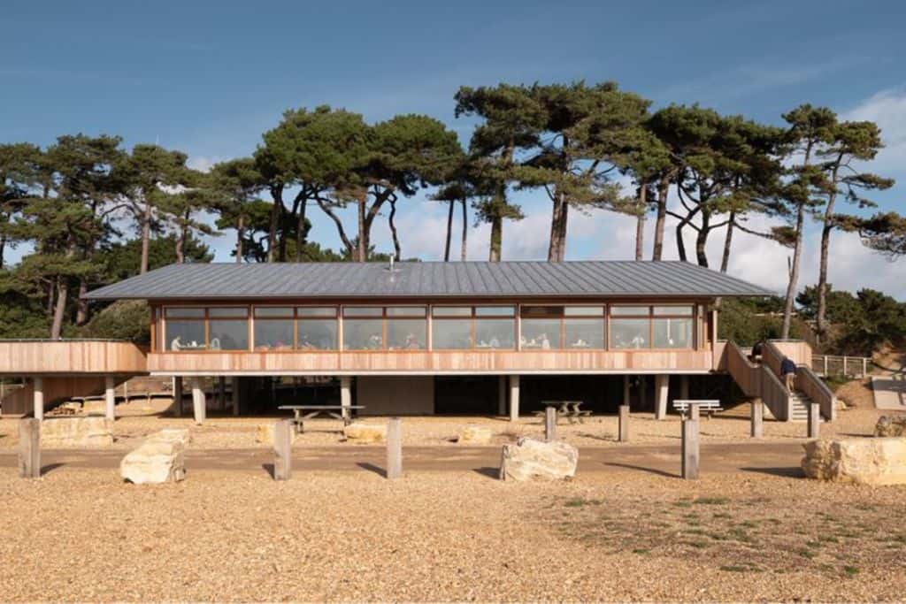 The outside of a wooden building on a shingle beach with some big trees behind it. This is the Lookout Cafe at Lepe Country Park which is one of the best things to do in Hampshire.