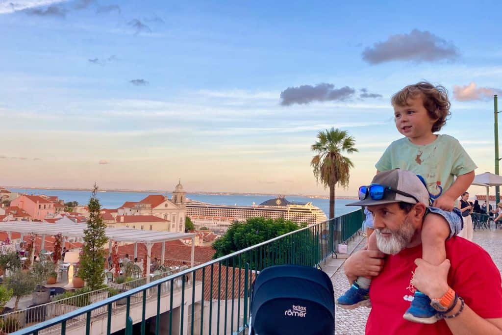 Father wearing red t-shirt with baseball cap with his young son on his shoulder looking out over the River Tagus in Portugal.