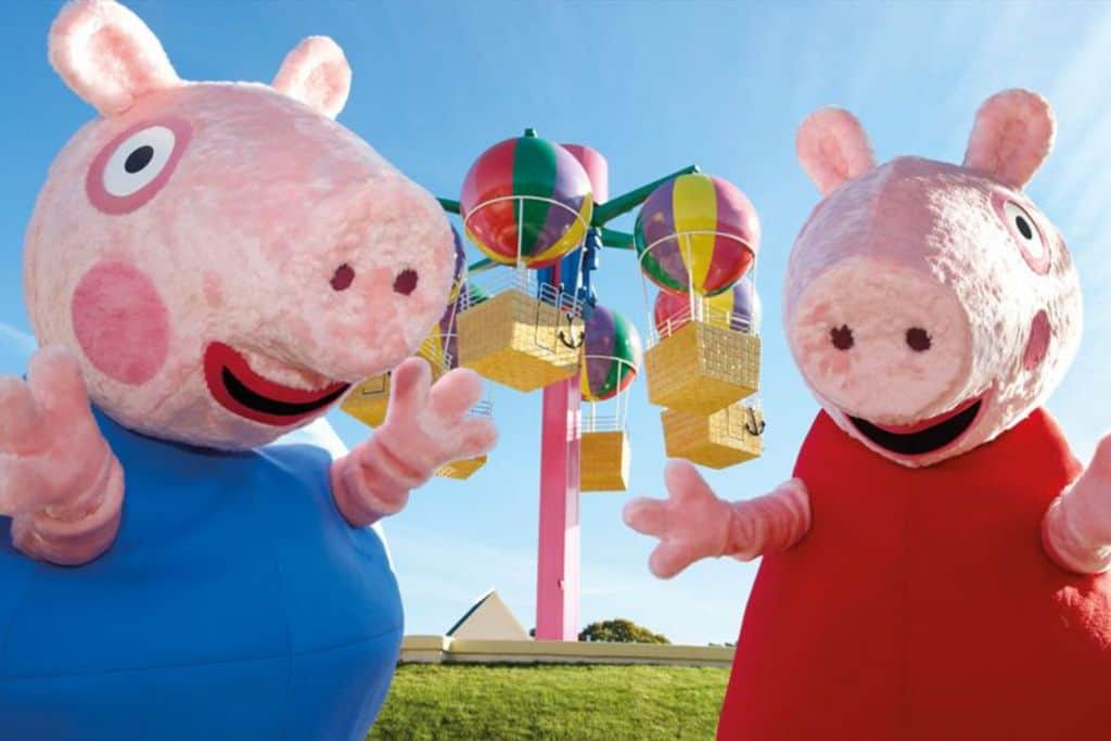 Two adults in fluffy Peppa and George Pig costumes in front of a fairgrounf ride at Paultons Park.  This is the only theme park childrens attractions in hampshire