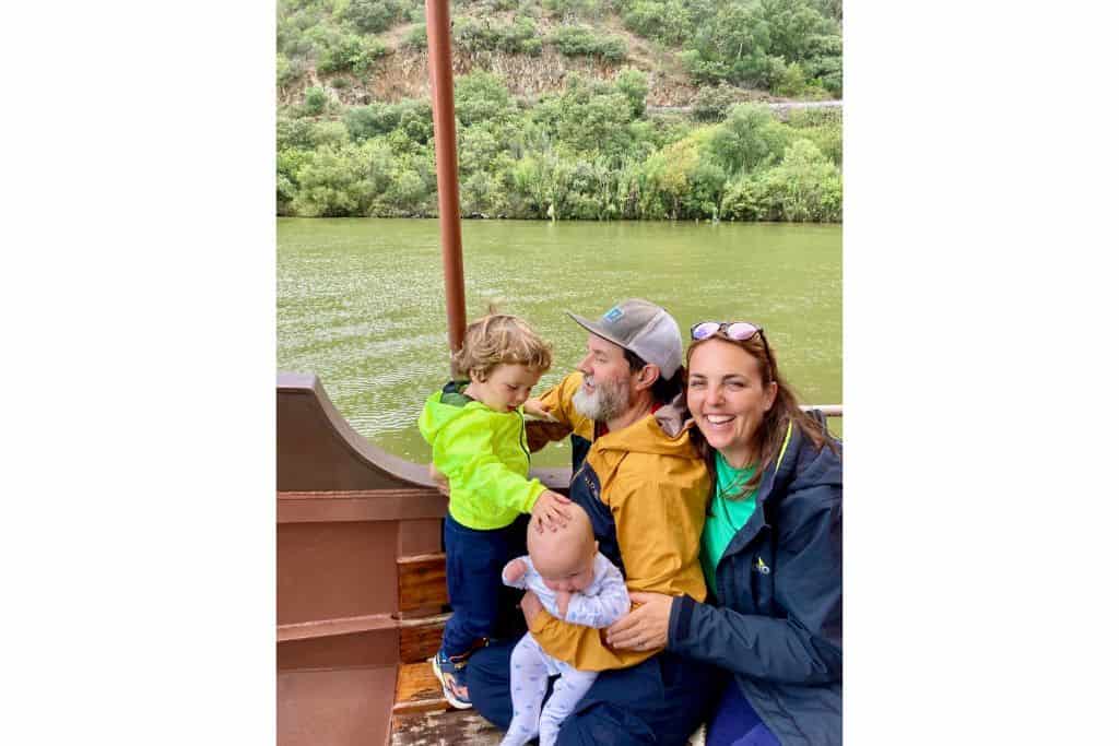 A mother, father and their two baby sons wearing rain jackets on a boat on the Douro River in Porto. Either side of the river are the vineyards.