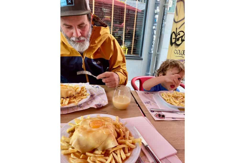 A father and son sat next to each other outside at a restaurant table eating a Francesinha sandwich which is a local dish in Porto.