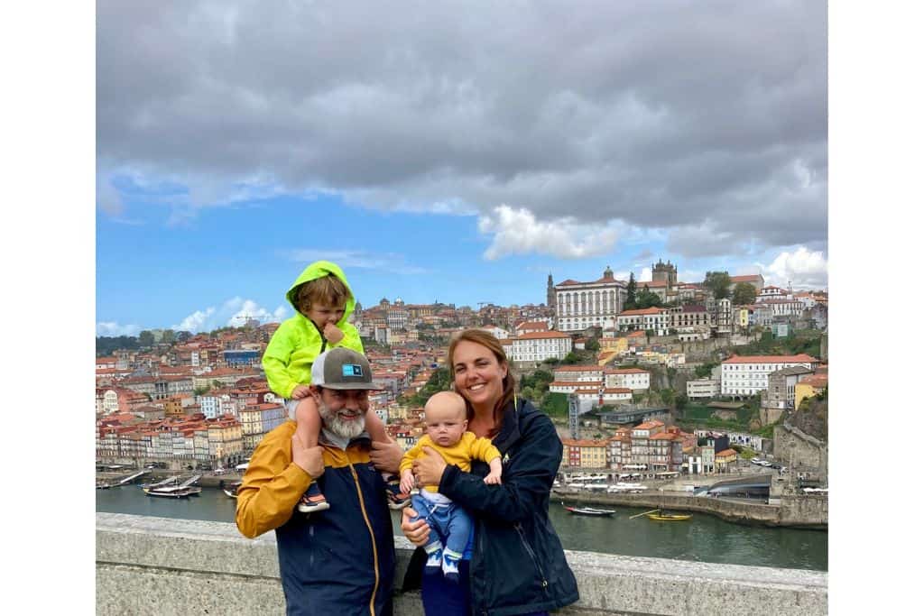 A dad in his raincoat with his young son sat on his shoulders.  Next to him is the mum holding their baby in her arms.  In the background is Porto.