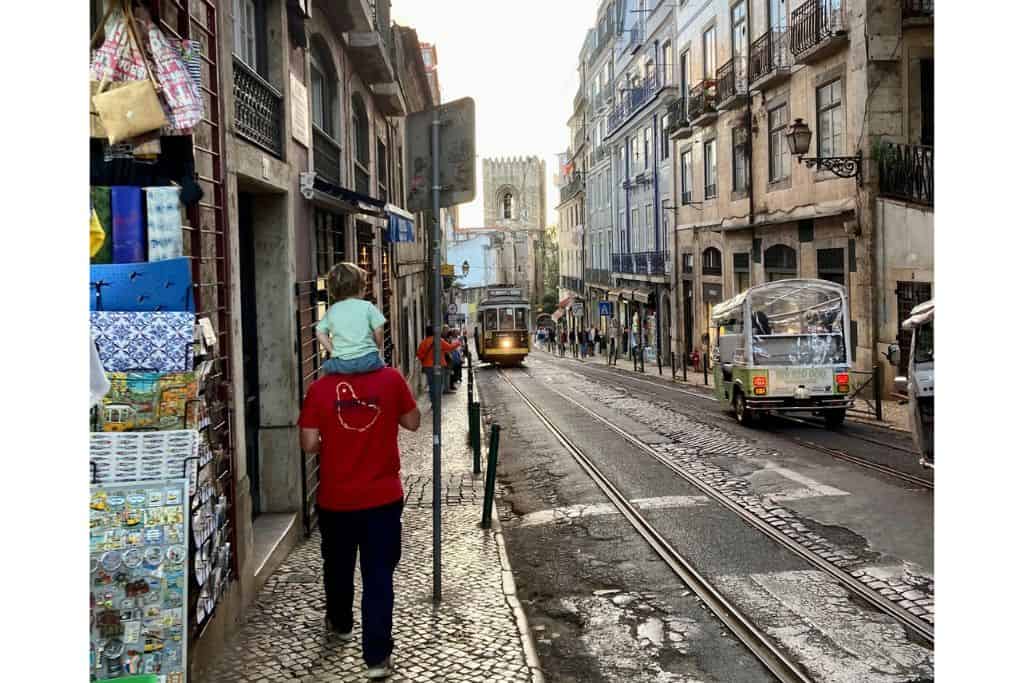 Man in a red t-shirt with his back to us with a young boy on his shoulders.  They are walking down a street in Lisbon with two trams on the street about to cross paths.