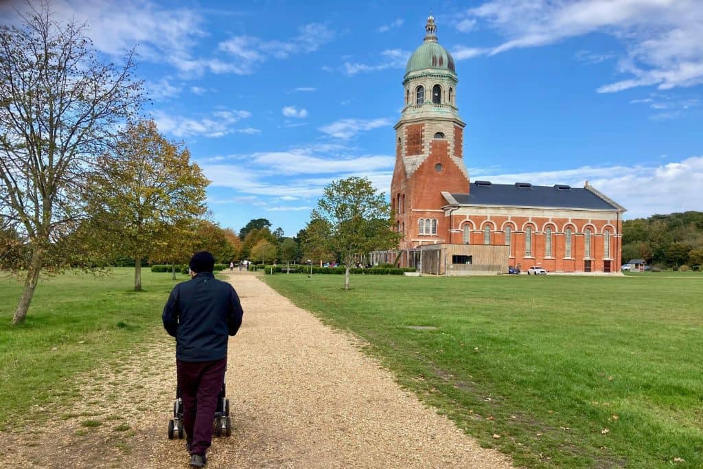 Father in a blue jacket pushing a pram along a path towards a chapel in Royal Victoria Country Park which is a fun childrens attractions in Hampshire.