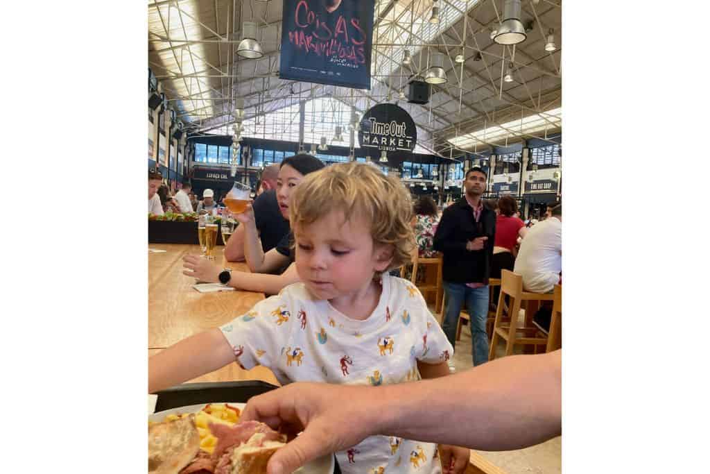 Young boy with animals on his t-shirt sat at a long communal high table.  He is eating and a sign in the background reads Time Out.  This is the Time Out foodhall in Lisbon which is one of the tun things to do in Lisbon with kids.