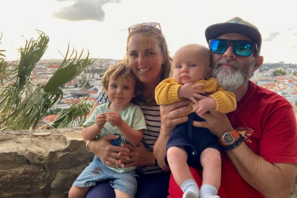 Family of four with includes a mum, dad, toddler and baby all sat on the wall of Lisbon Castle with the city view in the background.