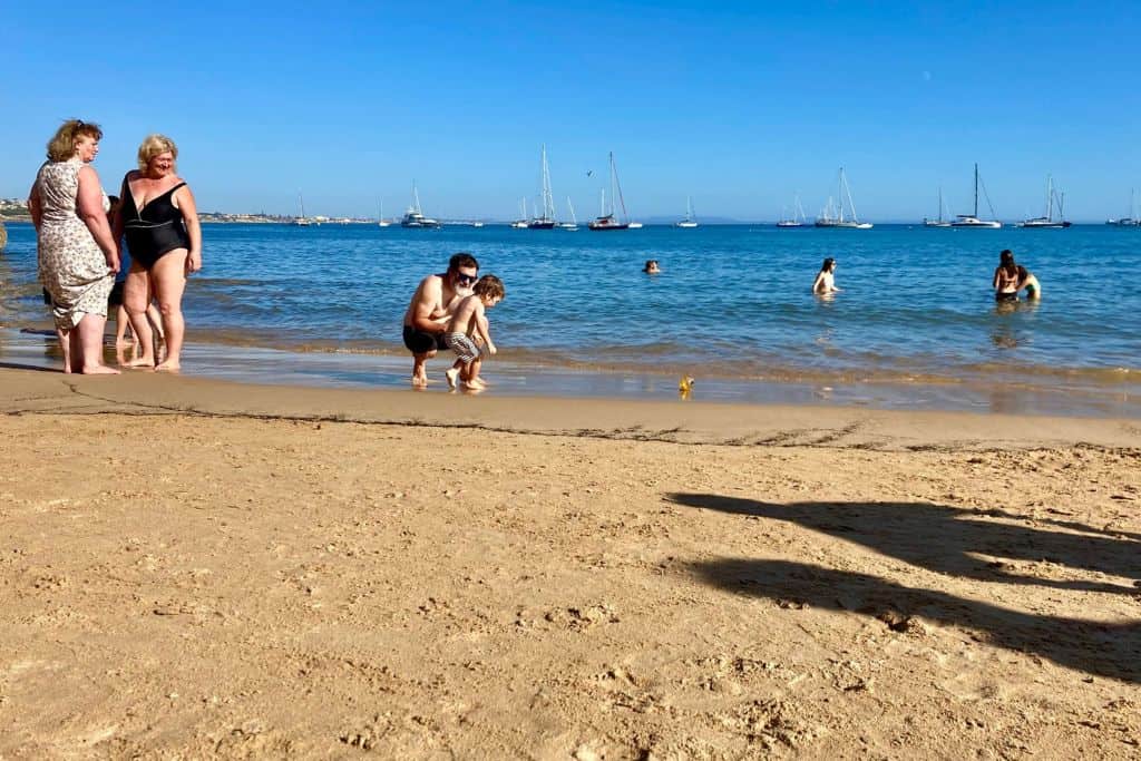 Golden sand beach with blue sea in the background.  On the beach which is in Lisbon are people walking around. In the middle of the image is a father kneeling down chatting to his son.