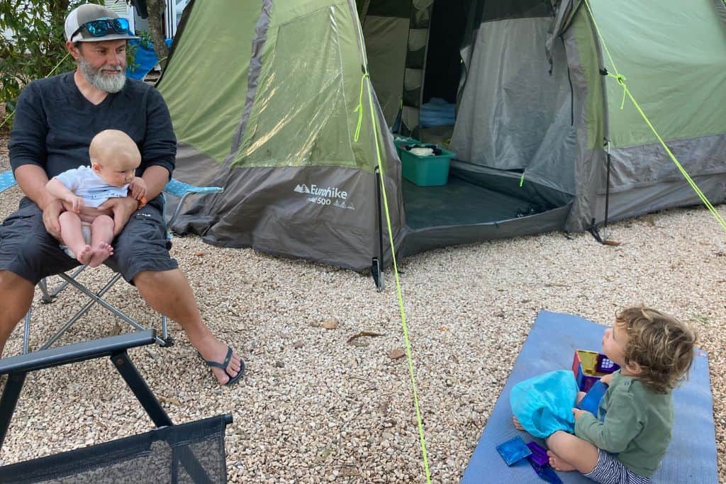 A father is sat on a chair with his baby son on his lap and his toddler son is sat on the floor.  In the background is their green tent.