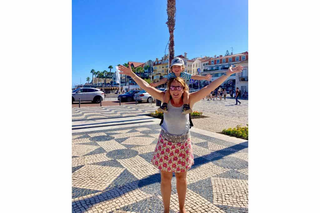 Mother in a grey vest top and mini skirt with flowers on it is stood in the centre of the main square in Caiscais with her toddler son on her shoulders.  They both have their hands in the air!