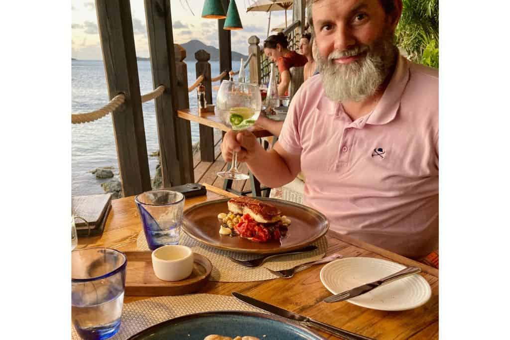 A man holding a gin and tonic in his hand in Sheer Rocks Restaurant in Antigua.  On the table is his main meal, a ceramic bowl of butter, extra cutlery as you'd expect in a luxury meal setting.