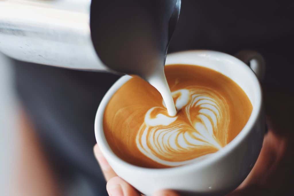 A jug containing hot milk is pouring milk into a freshly made coffee with coffee art on the top of the coffee.