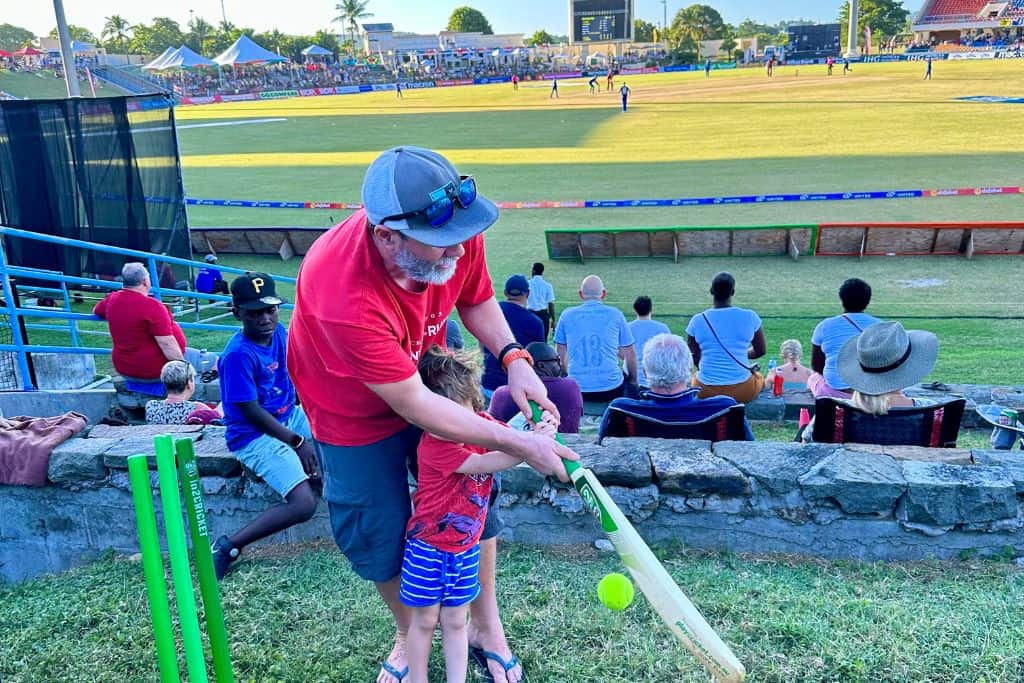 A father and son batting in a cricket match that is being played in the cricket stadium in Antigua whilst the actual match plays in the background.  You can visit the cricket ground from the Antigua cruise port.