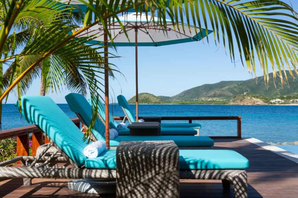 Three sun loungers on the terrace about the beach at Curtain Bluff Resort in Antigua.  Above the loungers are blue parasols. In the background is the sea with lush green mountains coming up from one side. 