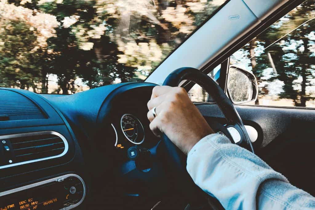 Inside of a car with someone's hands on the steering wheel.  From the windscreen you can see the passing trees that they are driving past.  Driving is one of the ways that you can get from Porto to Lisbon for a day trip.