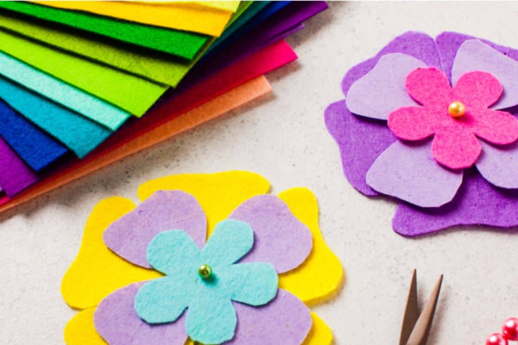 In the top left are a seleciton of pieces of different colour felt.  In the centre of the image is the starting point of two felt flowers, on the right there is one that it pink and purple, on the left is one that is yellow, purple and blue.