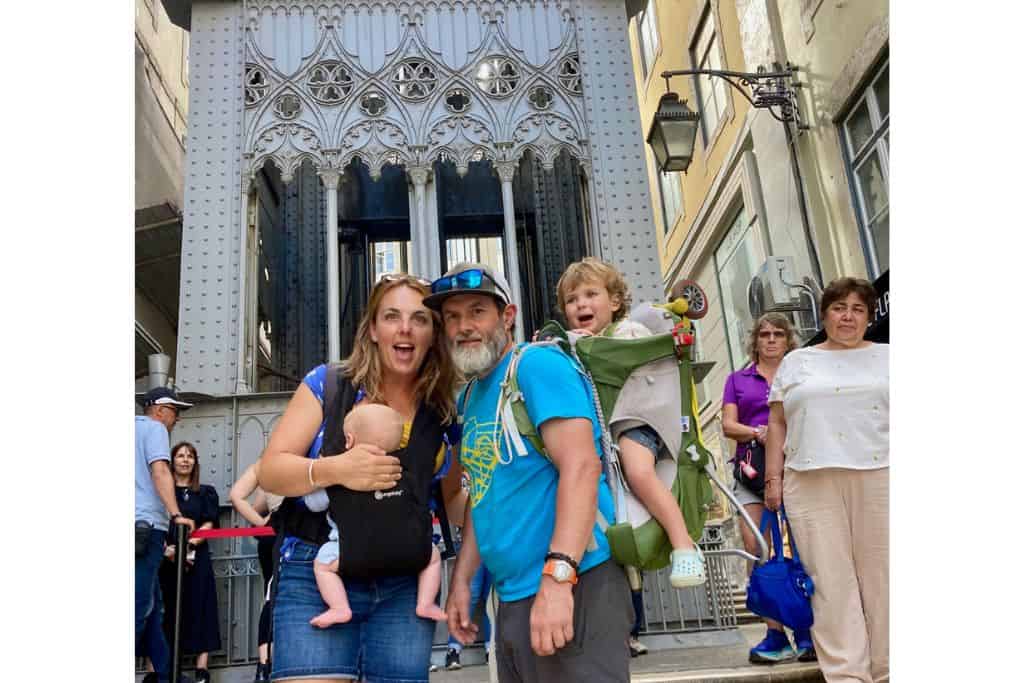 Family stood in from of a funicular. There is mum smiling with a baby in a carrier on her front. And dad in a bright blue t-shirt with a green child carrying backpack on his back with a young boy in it.  Riding a funicular in Lisbon is one of the things to do with kids.