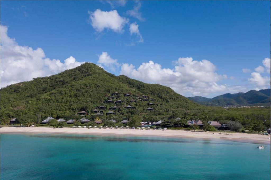 A view from the water looking back at the beach at Hermitage Bay Resort.  There is a lush green mountain and in front of it a beach with villas nestled above. There are also sun loungers and parasols on the beach.