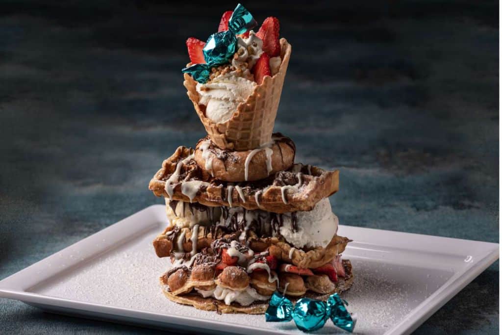 A dessert is on a white plate.  It is made up of waffles stacked on top of each other wtih ice crem sandwiched between them with cream on top and waffle ice cream cone with more ice cream in it.  All dripping with chocolate and caramel sauce.