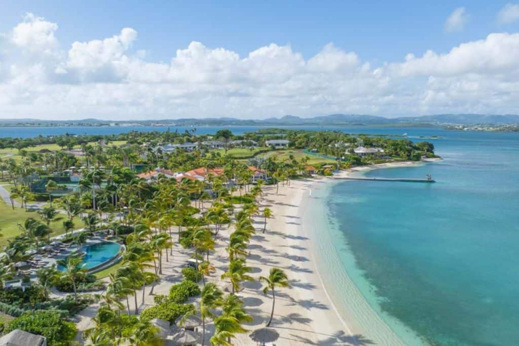 A view over Jumby Bay Resort on Long Island with trees and green gardens over the beach. The beach has white sands and the sea is crystal clear.  Jumby Bay is the most exclusive of the Antigua all inclusive luxury resorts.