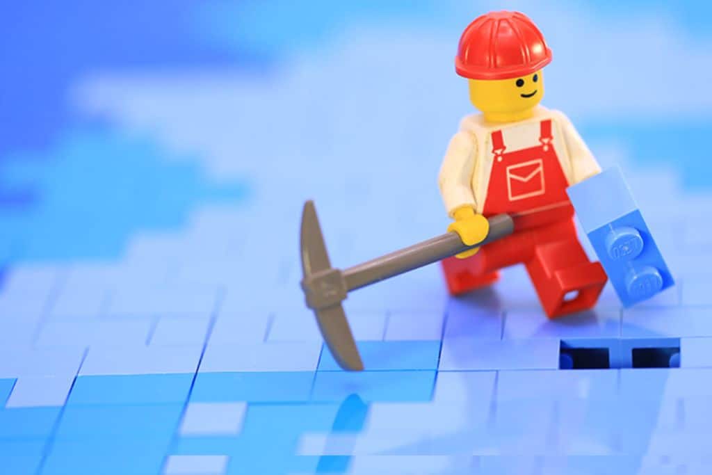 In the background is a floor mail of lego brick. In the centre right is a lego man in white shirt and red dungeress holding a Lego brink and a pick axd.