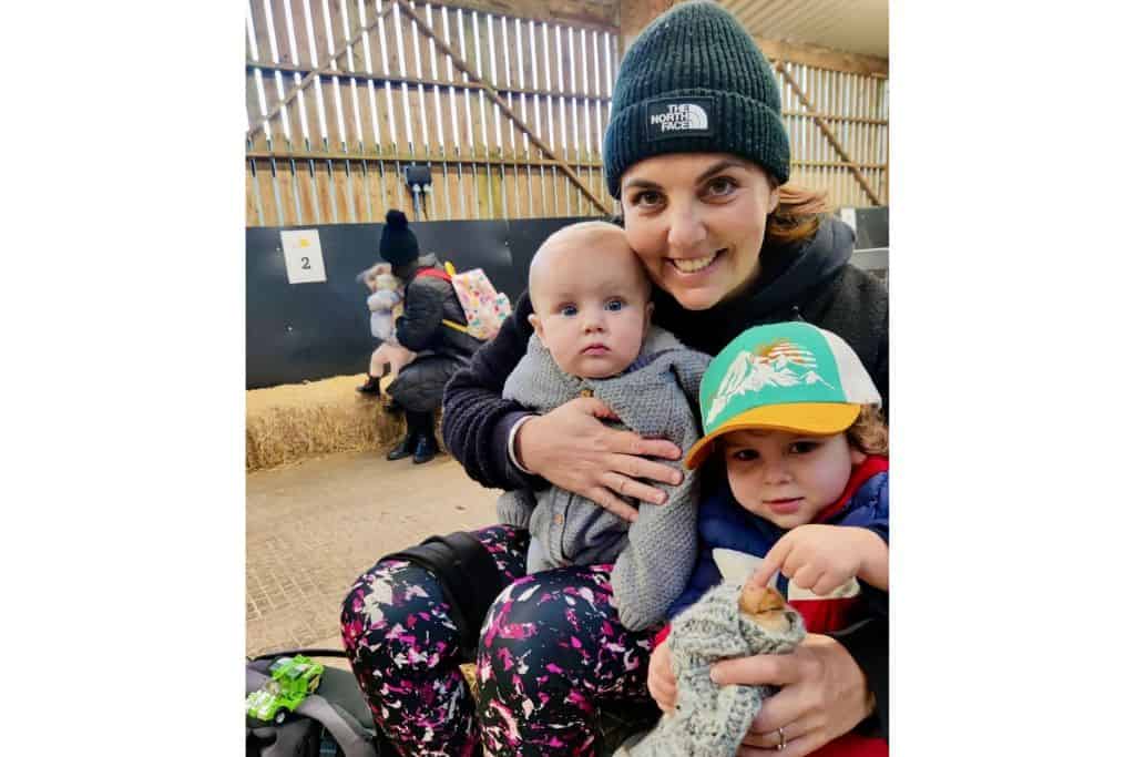 A mum and her two sons are sat on a hale bale in. barn.  The baby is in her arms in a grey onsie looking surprised.  The three year old is wearing a baseball cap and in his hands is a baby chick wrapped in a scarf.