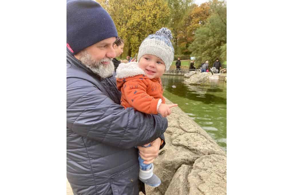 A father who is wearing a blue coat and beanie hat is holding his son who is wearing an orange coat and a beanie too.  They are looking over at the penguin enclosure which is part of Marwall Zoo which is a popular childrens attractions in Hampshire.