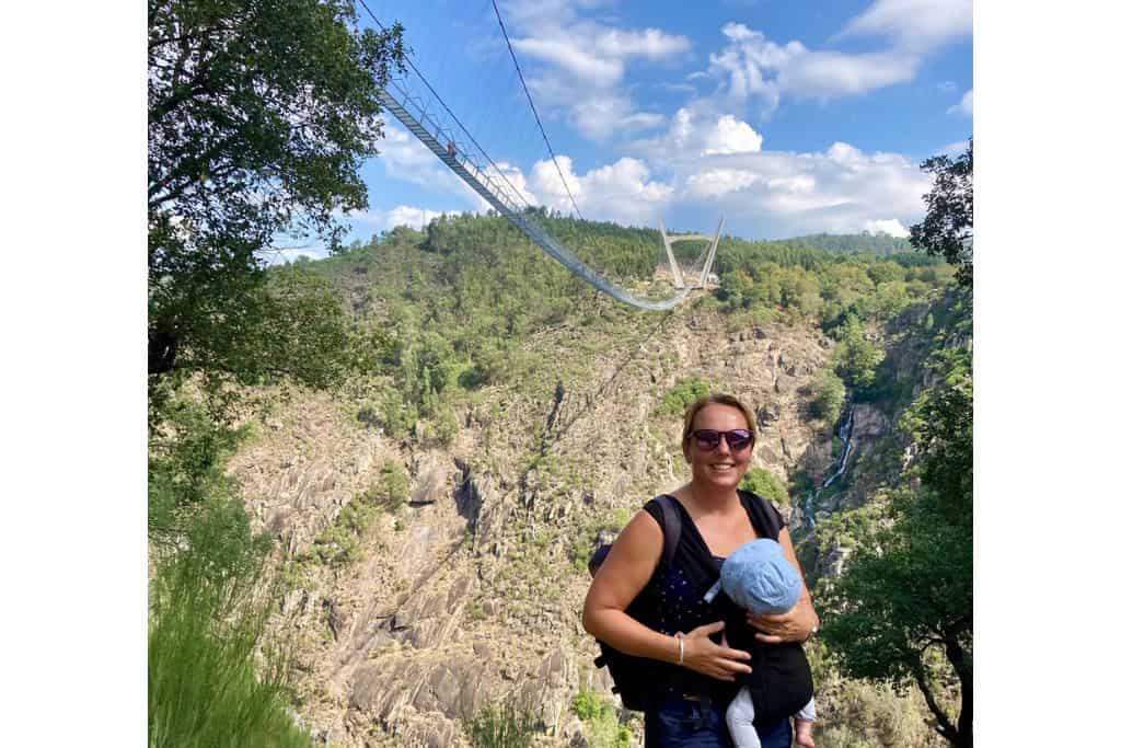 A mum with her baby in a carrier on her front is stood in front a pedestrian suspension bridge.  This long bridge is part of the Paiva Walkways near to Porto in Portugal and should go onto most peoples bucket lists.
