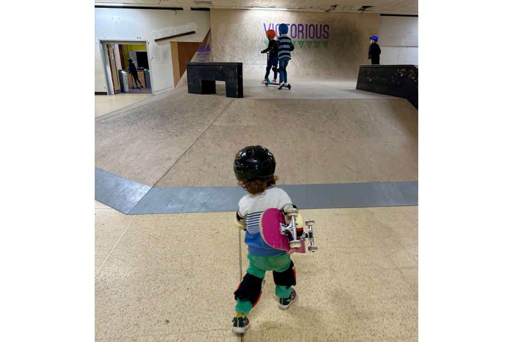 A young boy is walking across an indoor skate park with his pink skateboard under his arm. He is wearing a black helmet with black knee and elbow pads.  This is Pitt Street Skatepark which is a very popular childrens attractions in Hampshire.